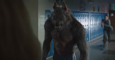 A large, furry werewolf with glowing eyes stands in a school hallway while a person runs in the background.
