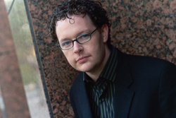 A man with dark curly hair and glasses wearing a dark suit and striped shirt leans against a granite wall.