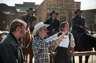 Men in cowboy hats and period clothing stand near horses in a dusty Western town.