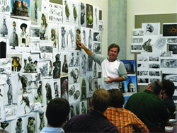 A man in a grey shirt points to character sketches on a wall covered in drawings.
