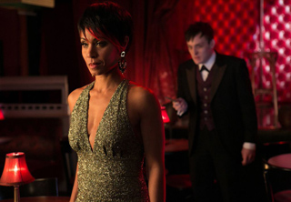 A woman in a sparkling green halter dress stands in a dimly lit room with a man in a tuxedo behind her.