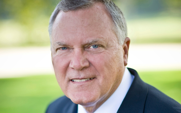 A close-up portrait of a middle-aged man with light hair and blue eyes wearing a dark suit and white shirt.