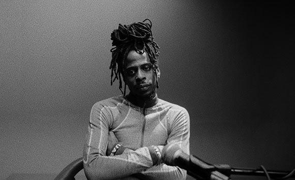 Black and white portrait of a man with long dreadlocks wearing a long-sleeved shirt and crossing his arms.
