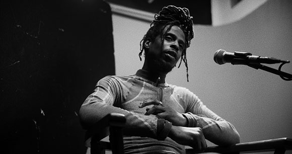 Black and white photo of a man with dreadlocks sitting behind a microphone.