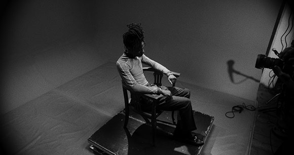 Black and white high-angle shot of a person with dreadlocks sitting in a wooden chair in a studio.