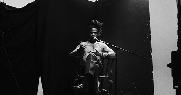 Black and white photo of a man with dreadlocks sitting in a chair and speaking into a microphone.