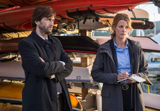 A man in a dark coat and a woman in a blue shirt and grey jacket stand before stacked kayaks.