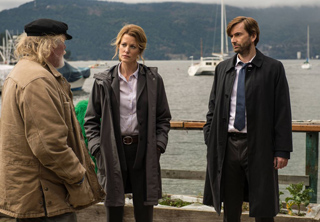 A woman and a man in dark coats face an older man in a tan jacket on a pier with boats in the background.