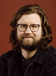 A man with wavy brown hair, a beard, and glasses looking forward against a reddish-brown background.