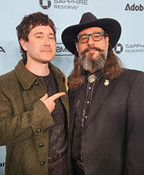 A man in a green coat points at a bearded man wearing a black hat and suit.