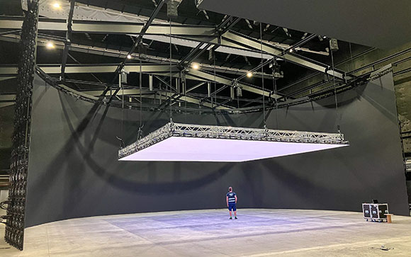 A person stands in a large dark studio featuring a curved LED wall and a large rectangular light panel overhead.