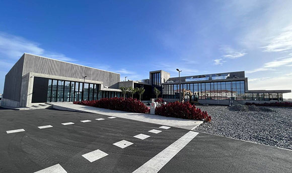 A modern, low-rise concrete building with large glass windows and red shrubbery under a clear blue sky.