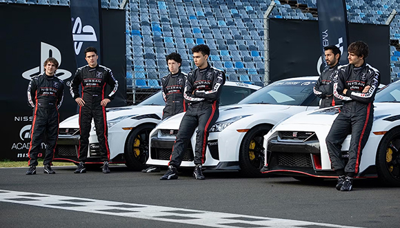 Five racing drivers in black and red suits pose with three white Nissan GT-R NISMO race cars.