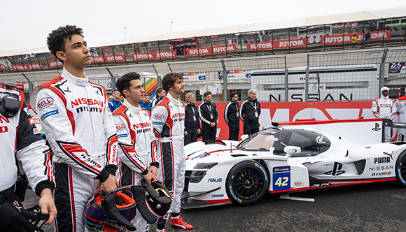 Racing drivers in Nissan NISMO uniforms stand next to a white and red Nissan race car on a track.
