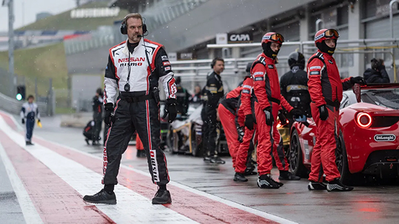 A man in a black and white Nissan Nismo racing suit stands on a wet track near crew in red suits.