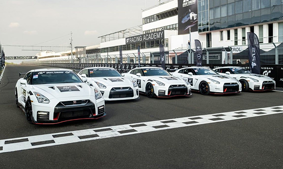 Five white Nissan GT-R race cars parked in a row on a racetrack in front of a racing academy building.