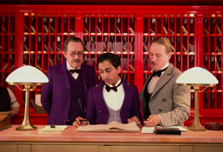Three men in vintage formal wear stand behind a counter in a red room with many small cubbies.