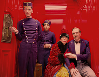 Four people in stylized purple and red uniforms and formal wear pose against a bright red wall.