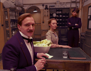 A man in a purple suit looks at the camera while a woman and a bellhop stand behind a counter.