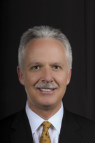 A smiling man with gray hair wearing a black suit, white shirt, and gold patterned tie against a black background.