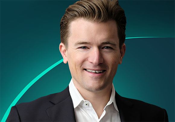A smiling man with brown hair wearing a black suit and white collared shirt against a teal background.