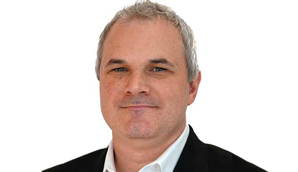 A man with short, graying hair, wearing a black suit jacket over a white shirt, looks directly at the camera.