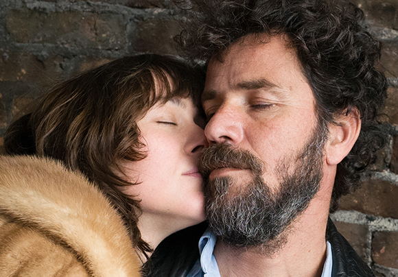 A woman with short brown hair closes her eyes while leaning her face against a bearded man's cheek.