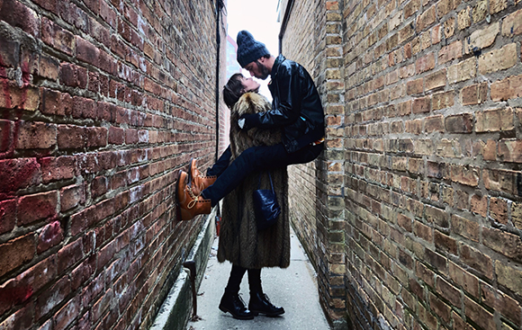 A man in a black jacket and beanie lifts a woman wearing a fur coat in a narrow brick alley.
