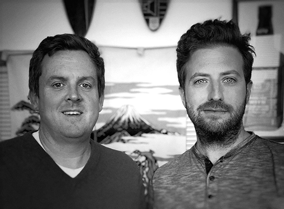 Black and white portrait of two men standing side by side in front of a landscape artwork.
