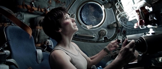 A woman in a tank top looks up through a circular spacecraft window at the Earth in space.