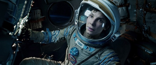 An astronaut in a white helmet and grey spacesuit looks intently at controls inside a dark spacecraft.