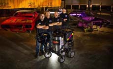 Four men pose in a garage with a red car, a purple car, and a piece of machinery.