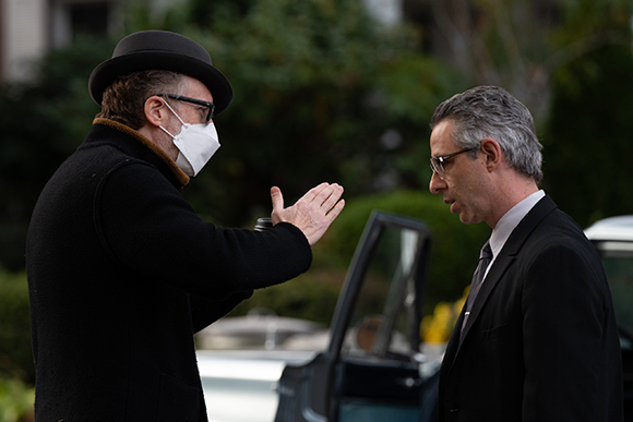 A man in a bowler hat and white mask gestures toward a man in a suit and glasses.