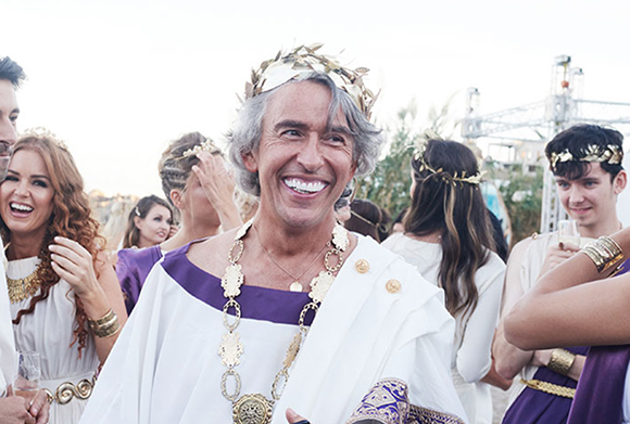 A smiling man in a white and purple toga and gold laurel wreath at an outdoor gathering.