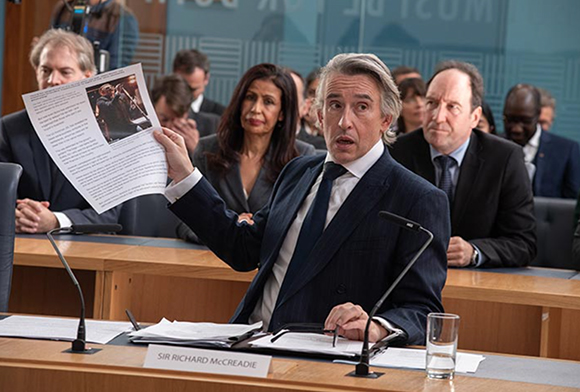 A man in a dark suit holds up a printed document featuring a photograph while seated at a conference table.