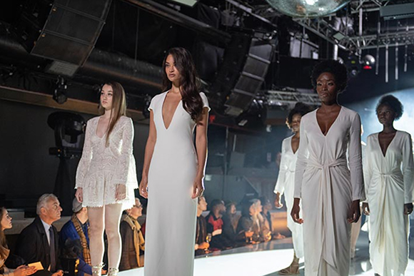 Models in white gowns walk down a brightly lit fashion runway during a show.