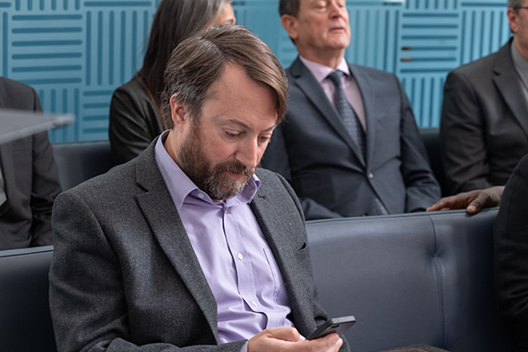 A man in a grey blazer and lavender shirt looks down at a smartphone while seated in a blue lounge.