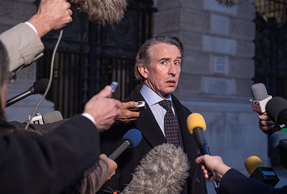 A man in a dark coat and tie speaks into multiple microphones held by reporters.