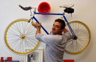A man in a grey shirt poses behind a blue bicycle mounted high on a white wall.
