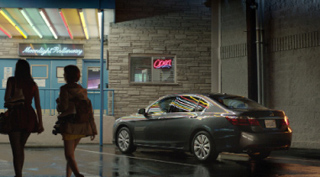 Two women walk past a motel with neon signs and a parked dark sedan at night.