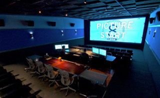 A dark screening room featuring a large screen displaying 'PICTURE START' and a control desk with monitors.