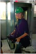 A woman wearing a green hard hat, safety glasses, and a dark shirt stands in a doorway.