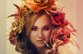 A blonde woman wearing a large crown of orange and red flowers, holding a cigarette in her mouth.