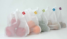 Four white mesh reusable bags containing different fruits and vegetables lined up on a white surface.