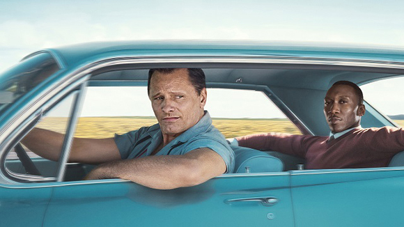 Two men sit in a light blue vintage car against a landscape of golden fields.