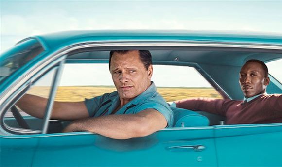 A man drives a blue vintage car while another man sits in the passenger seat looking forward.