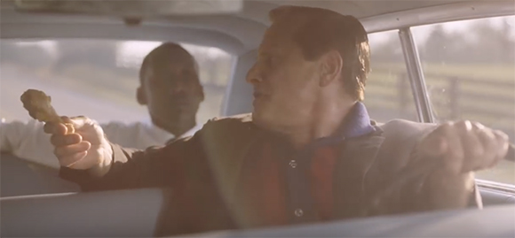 A man in a car holds up a piece of fried chicken while looking toward another man in the backseat.