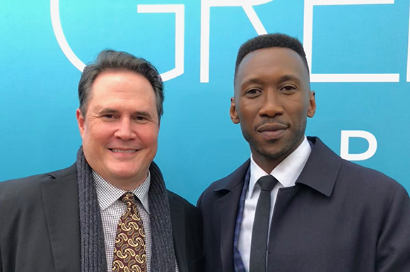 A man in a suit and scarf standing next to a man in a dark coat and tie against a blue backdrop.