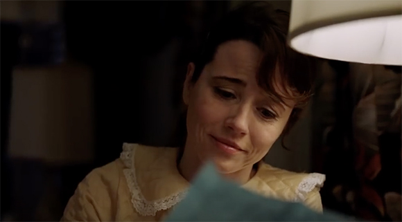 A woman with dark hair looks down with a sorrowful, tearful expression in a dimly lit room.