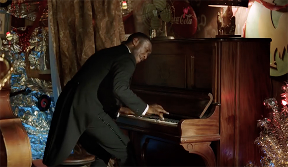 A man in a black suit leans forward while playing the keys of a dark wood upright piano.